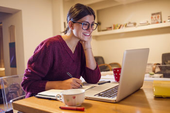 Junge Studieninteressentin sitzt Zuhause am Schreibtisch vor einem Laptop und notiert sich Zulassungsvoraussetzungen