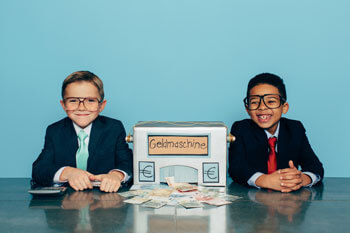 Zwei kleine Jungen in Anzug und Brille sitzen neben einer gebastelten Geldmaschine
